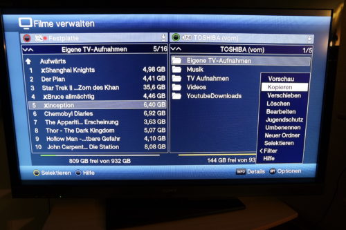 Im Menü Filme-Verwalten ist der Bildschirm zweigeteilt. Auf der einen Seite wird der Inhalt der internen Platte angezeigt, auf der anderen Seite der Inhalt des angestöpselten Mediums. Ein Klick auf OPT öffnet ein weiteres Menü. Hat man mehrere Filme zu kopieren, wählt man "Selektieren" und wählt dann mit der gelben Taste die gewünschten Filme aus. Anschließend eion neuer Druck auf OPT und dann wählt man KOPIEREN und schon startet der Kopiervorgang, der sich über einige Zeit hinziehen kann.